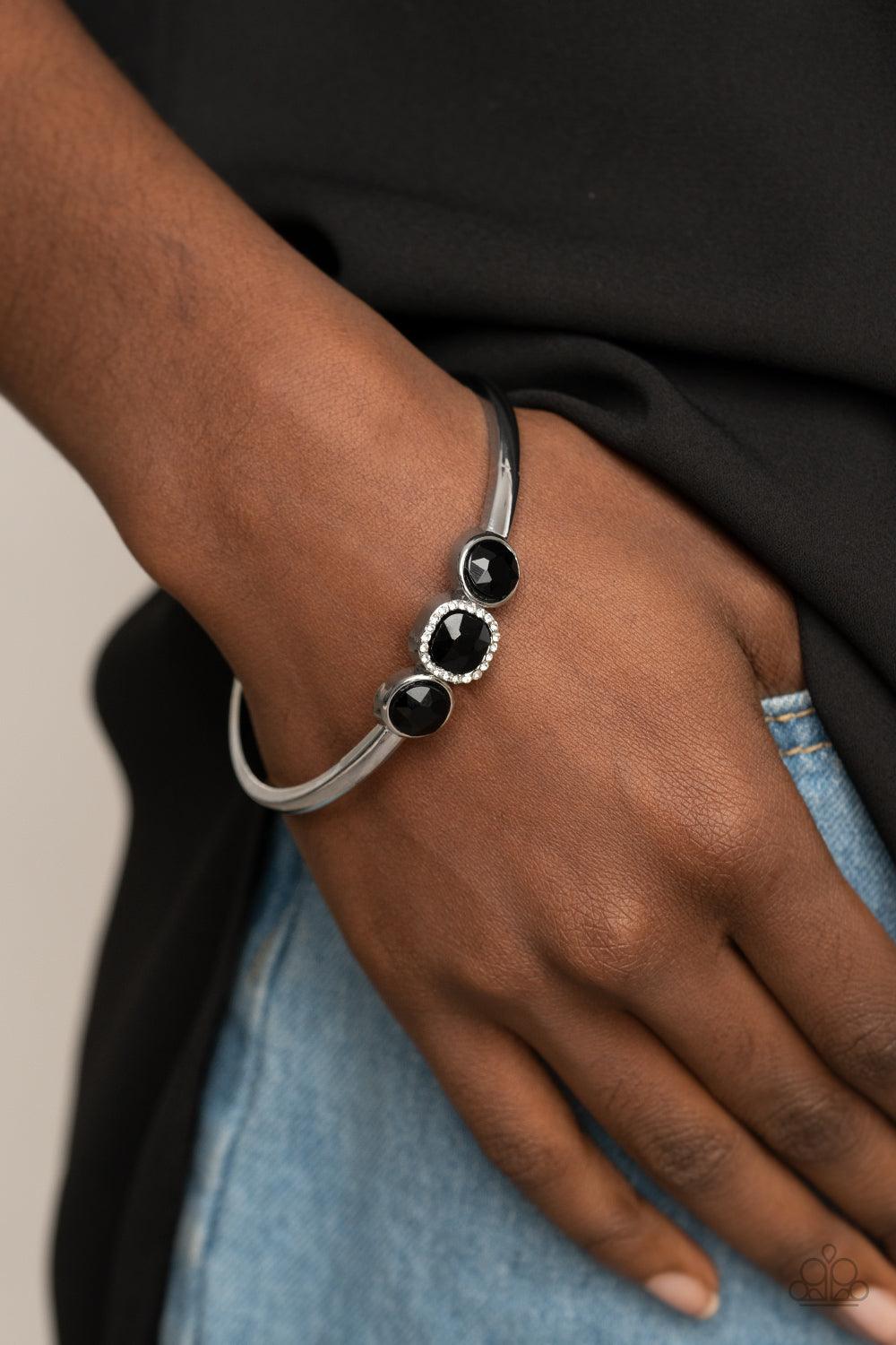 Paparazzi Accessories Royal Demands - Black The center of a thick silver bangle has been adorned with a trio of glittery black rhinestones, creating a sparkly centerpiece around the wrist. Featuring a regal cube cut, the centermost rhinestone is bordered
