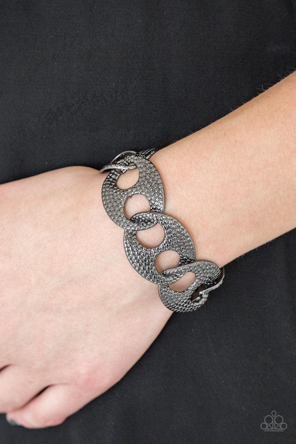 Paparazzi Accessories Casual Connoisseur - Black Embossed in shimmery circular patterns, asymmetrical gunmetal frames link across the wrist for a casually industrial look. Features an adjustable clasp closure. Jewelry